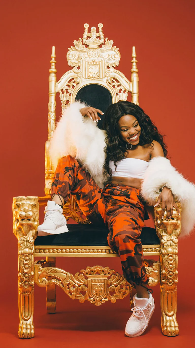 Lady Leshurr sits relaxed and smiling on a golden throne against a red background.