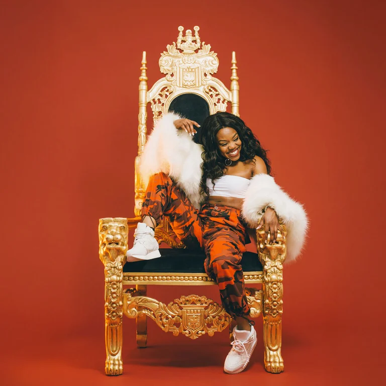 Lady Leshurr sitzt entspannt und ächeldn auf einem goldenen Thron vor rotem Hintergrund.