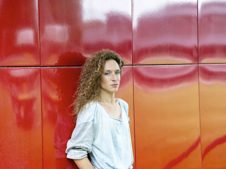 Francois Chiagnaud leans against a shiny red wall. They wear a loose, light-colored top and look into the camera with a serious expression.