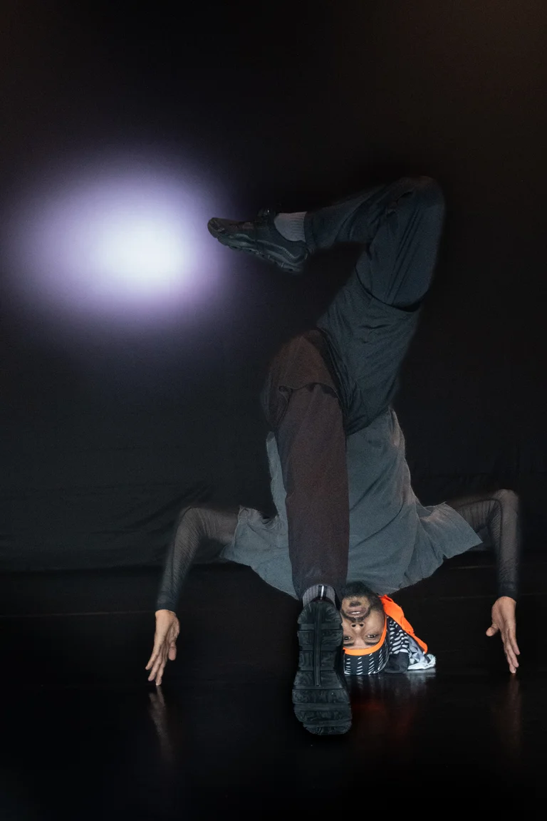 Mohamed Toukabri befindet sich in einer dynamischen, kopfüber ausgeführten Bewegung,  ein Bein gestreckt in der Luft, die Arme weit auf dem Boden abgestützt. Er trägt eine orangefarbene Kopfbedeckung, sein Gesicht ist ausdrucksstark zur Kamera gewandt, im Hintergrund leuchtet ein kreisförmiger Lichtschein auf dunkler Bühne.