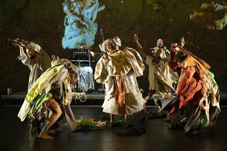 Dancing performers in colorful layered costumes on stage, with musicians and projections in the background.