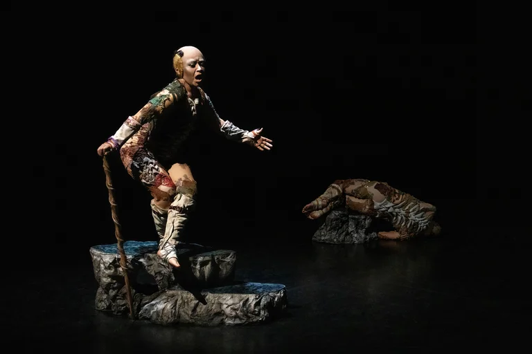 A woman stands on a rock-like platform holding a long staff. She wears a colorful, textured costume and appears to speak or call out in an expressive gesture. In the background, another person lies curled up on a smaller rock. The stage is dark.