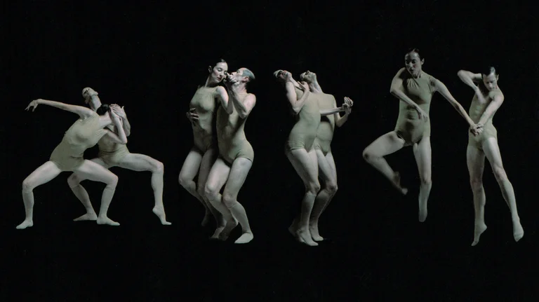Several dancers in skin-toned costumes move against a black background. The scene conveys tension, dynamism, and a strong sense of physical interaction.