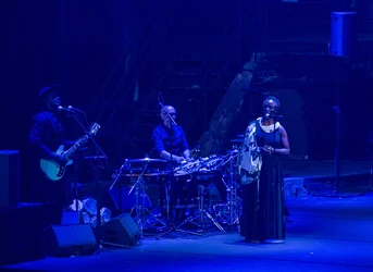 Three musicians perform on a blue-lit stage: a guitarist and a singer stand at microphones while a percussionist plays between them.