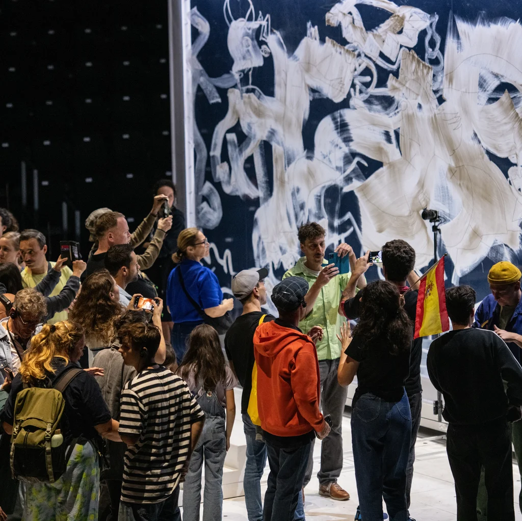 Eine Gruppe von Menschen versammelt sich vor einer großen Wand, die mit weißen Pinselstrichen und skizzierten Figuren bedeckt ist. Mehrere Menschen fotografieren die Szene, während jemand eine spanische Flagge hochhält.