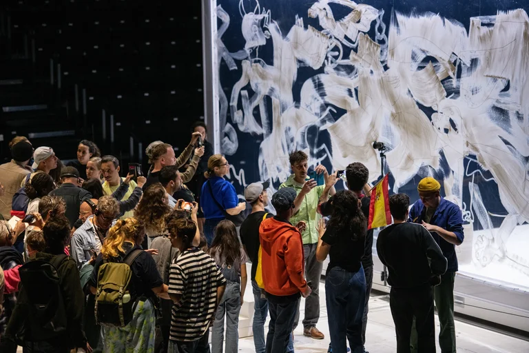 Eine Gruppe von Menschen versammelt sich vor einer großen Wand, die mit weißen Pinselstrichen und skizzierten Figuren bedeckt ist. Mehrere Menschen fotografieren die Szene, während jemand eine spanische Flagge hochhält.