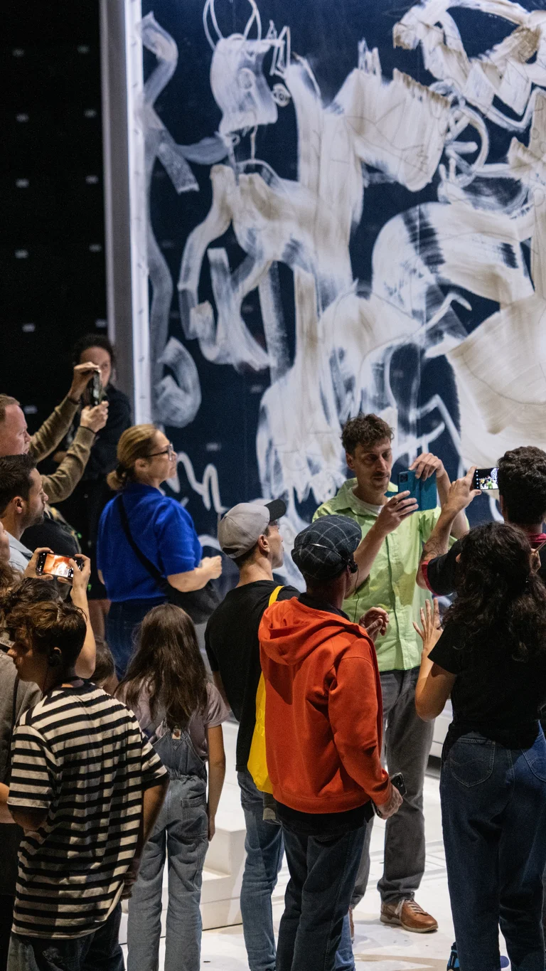 Eine Gruppe von Menschen versammelt sich vor einer großen Wand, die mit weißen Pinselstrichen und skizzierten Figuren bedeckt ist. Mehrere Menschen fotografieren die Szene, während jemand eine spanische Flagge hochhält.