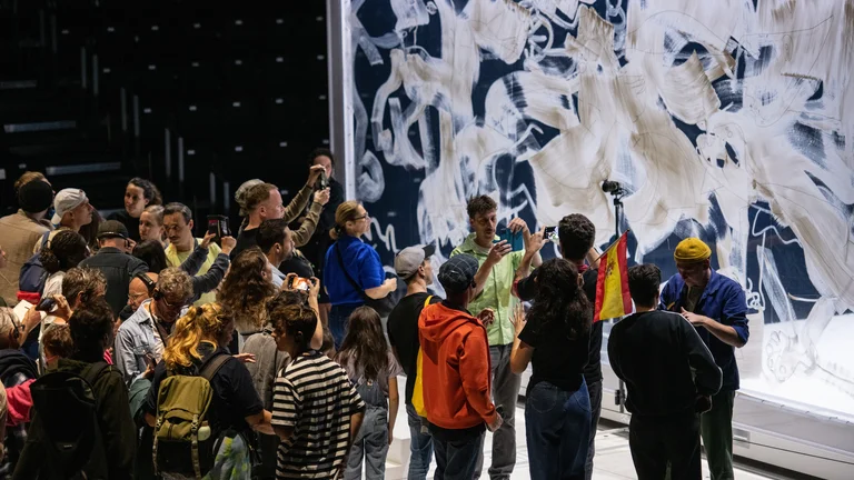 Eine Gruppe von Menschen versammelt sich vor einer großen Wand, die mit weißen Pinselstrichen und skizzierten Figuren bedeckt ist. Mehrere Menschen fotografieren die Szene, während jemand eine spanische Flagge hochhält.