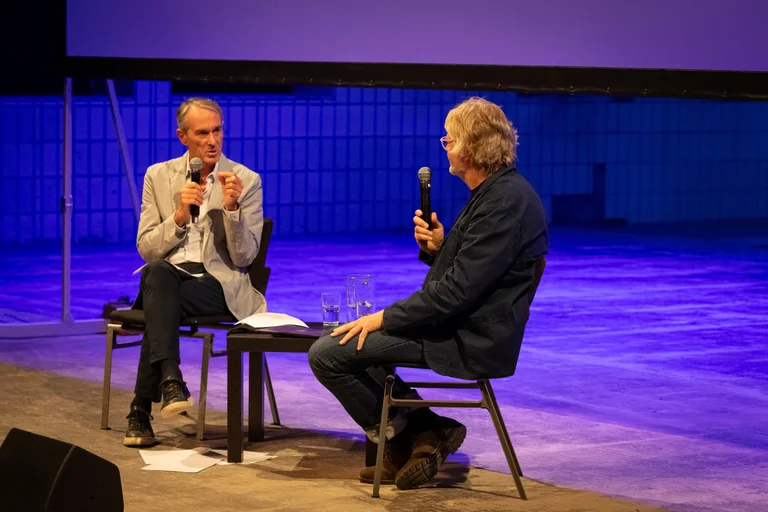 Ivo Van Hove and Will Gregory sitting on stage with microphones having a conversation.