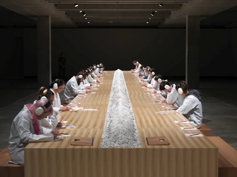 Participants sit at a long table counting grains of rice and lentils in Marina Abramović’s work “Counting the Rice”.