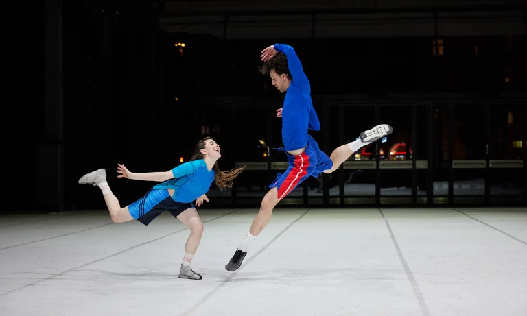 Zwei Tänzerinnen der Dresden Frankfurt Dance Company interagieren in bunten Kostümen auf einer Bühne.