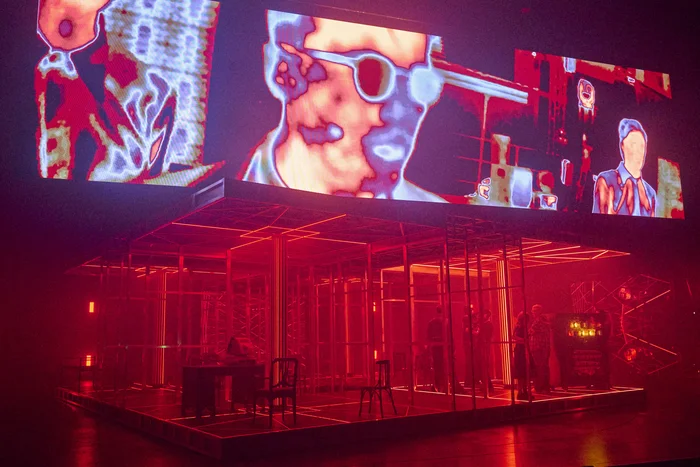 Stage scene in intense red lighting with a futuristic room structure; above it a projection of distorted video images.