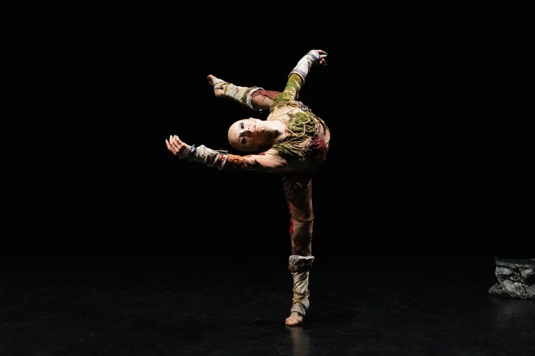 A person stands on one leg in a dynamic dance pose, with arms extended wide and one leg raised. They wear a colorful costume and gaze forward with an intense expression. The background is dark, making the figure stand out prominently.