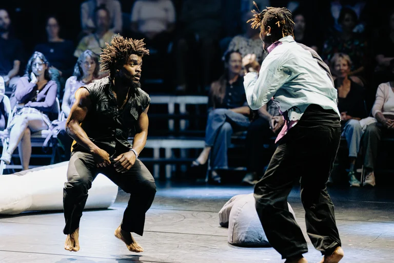 Two dancers are looking at each other. They are barefoot and dancing energetically. Only the back of the dancer on the right is visible; he is wearing a light-colored, stained shirt. The audience is seated in the background.