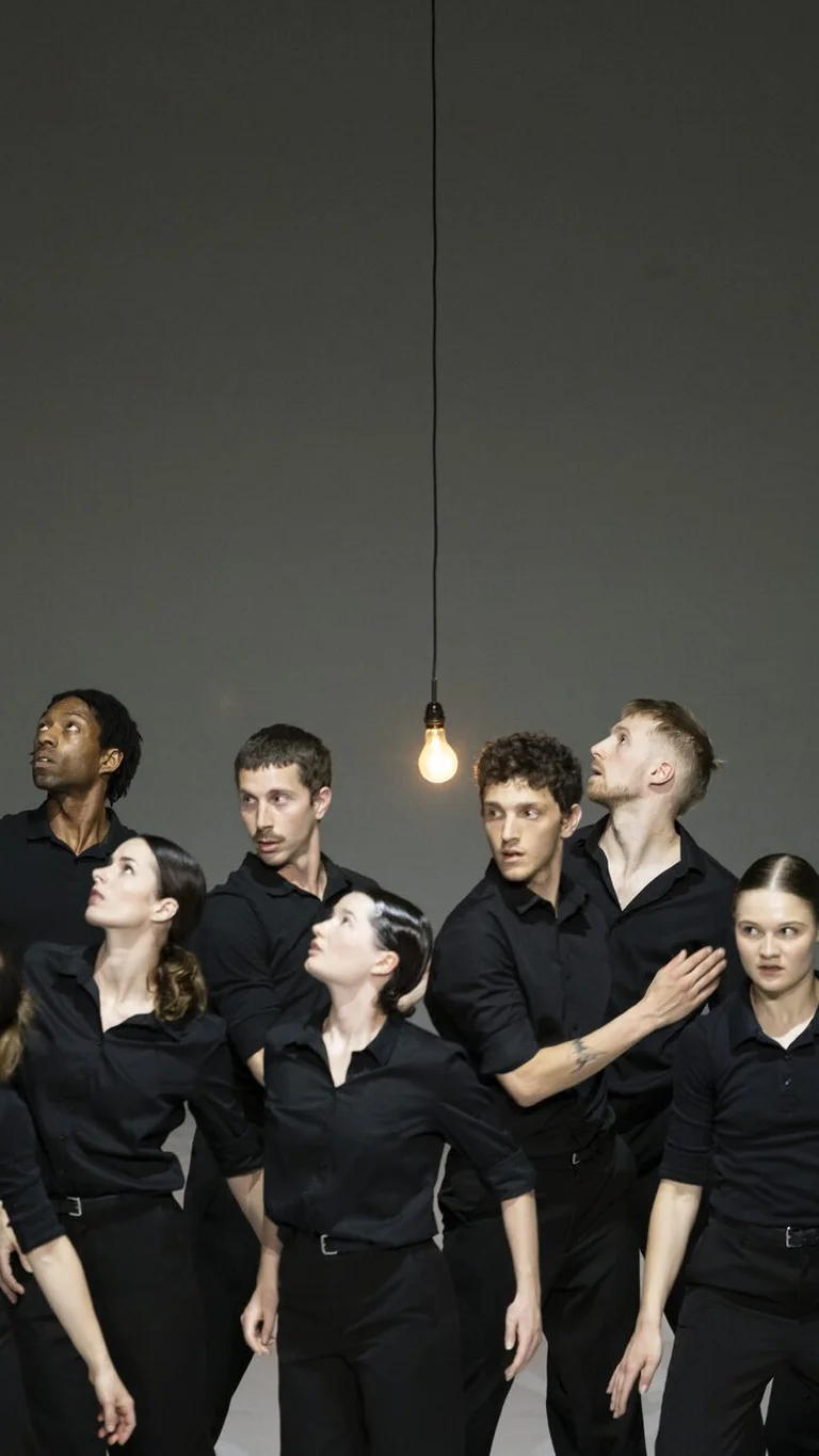 Group of performers in black clothing look up together at a hanging light bulb.