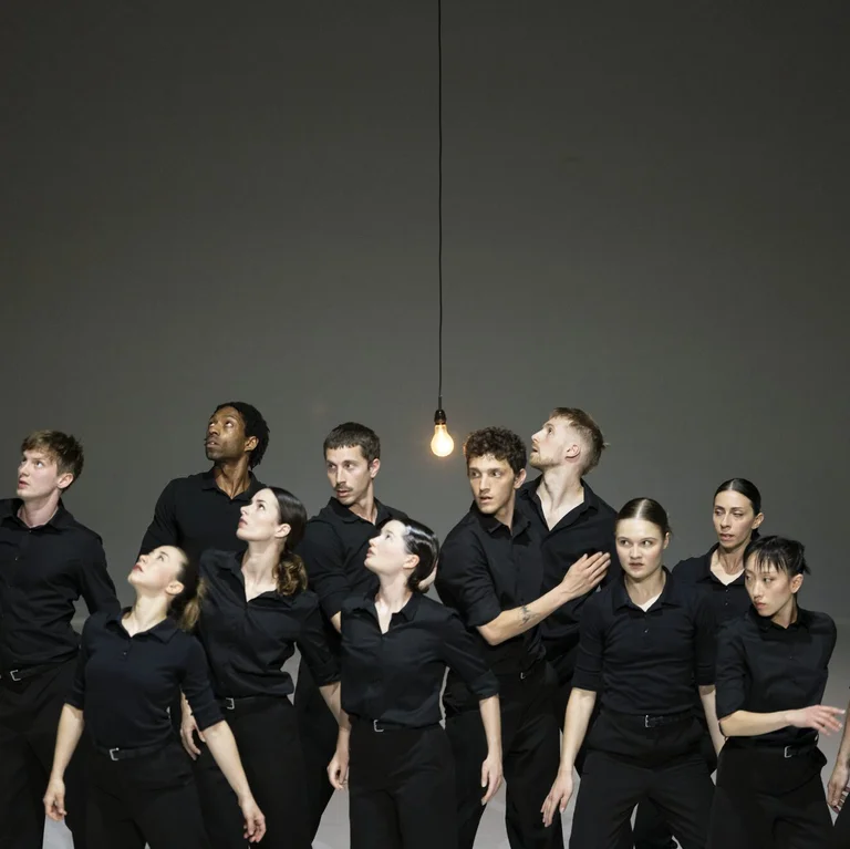 Gruppe von Performer:innen in schwarzer Kleidung blickt gemeinsam zu einer hängenden Glühbirne auf.