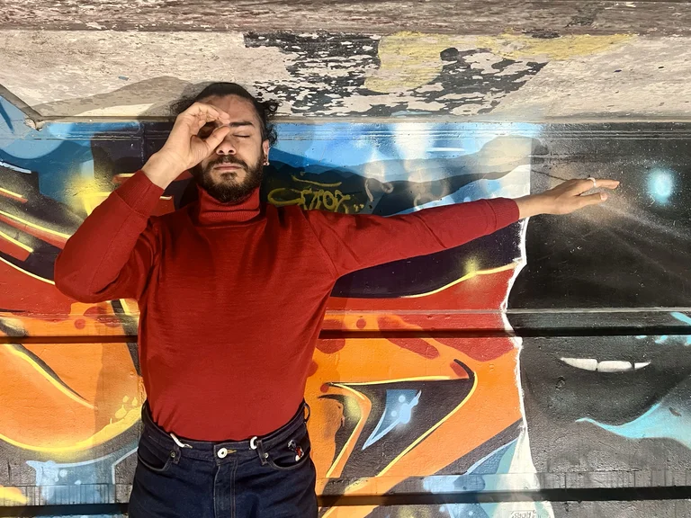 Mohamed Toukabri liegt auf dem Rücken vor einer bunten Graffiti-Wand. Er trägt einen roten Rollkragenpullover und dunkle Jeans. Sein rechter Arm ist ausgestreckt, während seine linke Hand ein Auge mit Daumen und Zeigefinger umkreist.