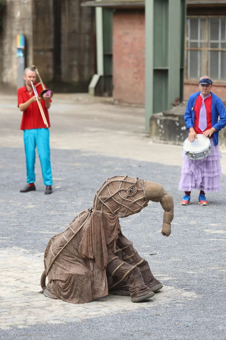 A small elephant sits on the floor. Musicians stand in the background and play a drum and a trumpet. The parade took place in Duisburg Landscape Park