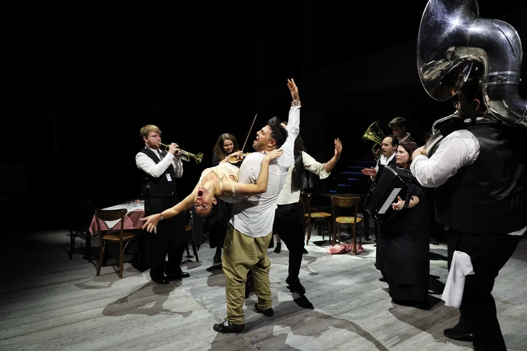 On a stage, a couple dances with the woman dramatically dipped back, surrounded by musicians with brass instruments, accordion, and a sousaphone.