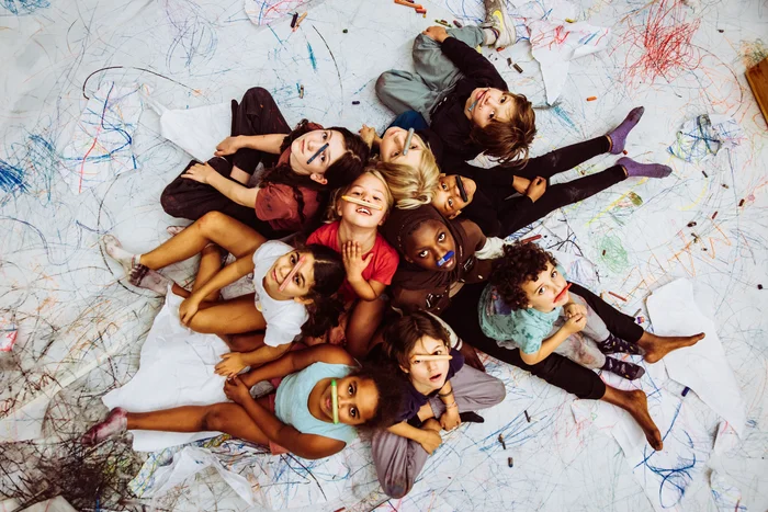 Eine Gruppe von Kindern liegt auf einem großen weißen Boden, der mit bunten Kritzeleien bedeckt ist. Sie blicken fröhlich nach oben zur Kamera und halten sich bunte Wachsmalstifte vor das Gesicht oder zwischen die Lippen.