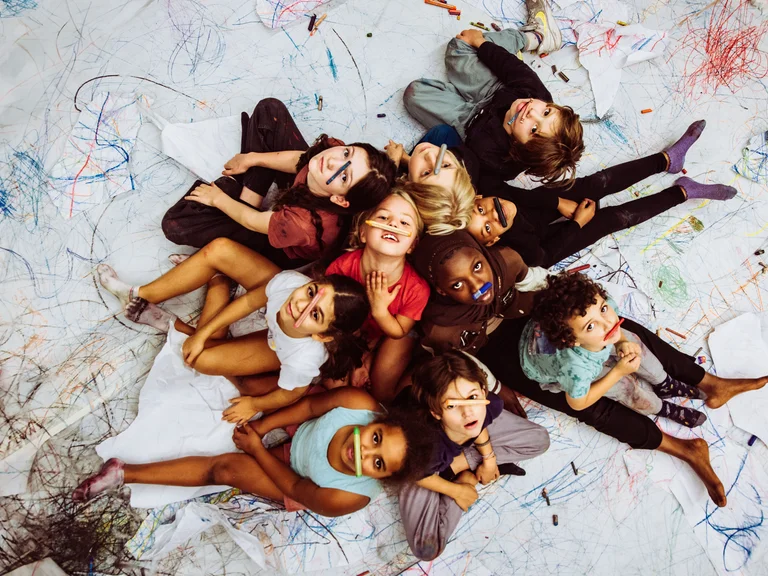 Eine Gruppe von Kindern liegt auf einem großen weißen Boden, der mit bunten Kritzeleien bedeckt ist. Sie blicken fröhlich nach oben zur Kamera und halten sich bunte Wachsmalstifte vor das Gesicht oder zwischen die Lippen.