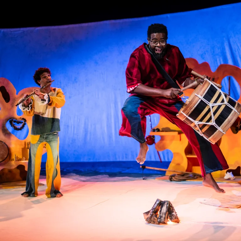 Three musicians play flute and percussion on a colourful stage as the drummer leaps into the air.