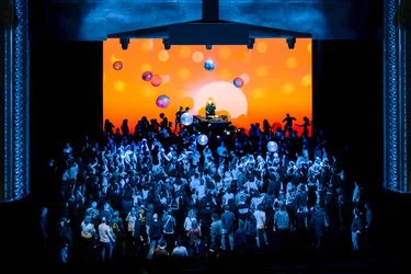 Eine große Menschenmenge tanzt vor einer leuchtend orangefarbenen Bühnenprojektion mit Discokugeln und Ballons, während ein DJ auf einem Podest auflegt.