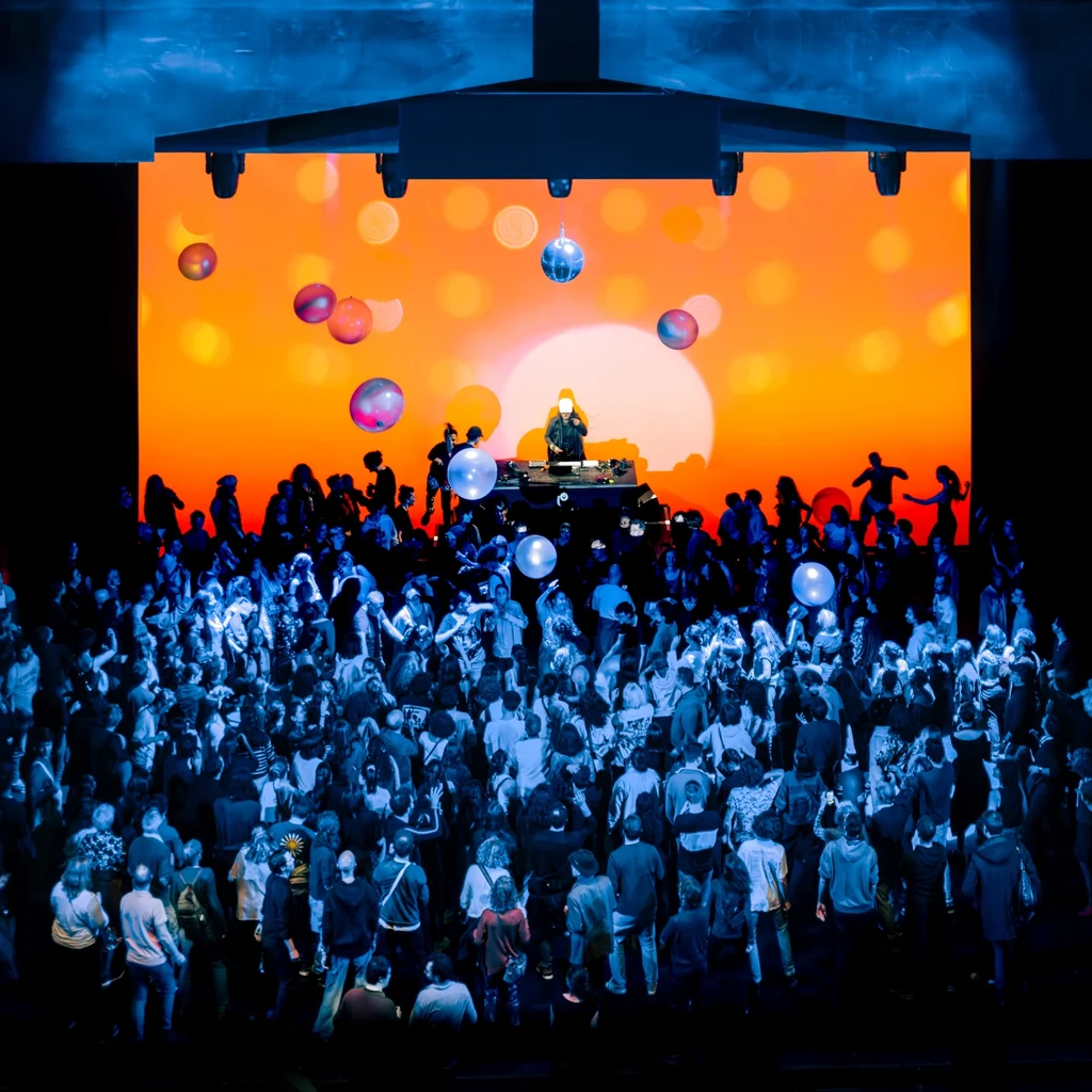Eine große Menschenmenge tanzt vor einer leuchtend orangefarbenen Bühnenprojektion mit Discokugeln und Ballons, während ein DJ auf einem Podest auflegt.