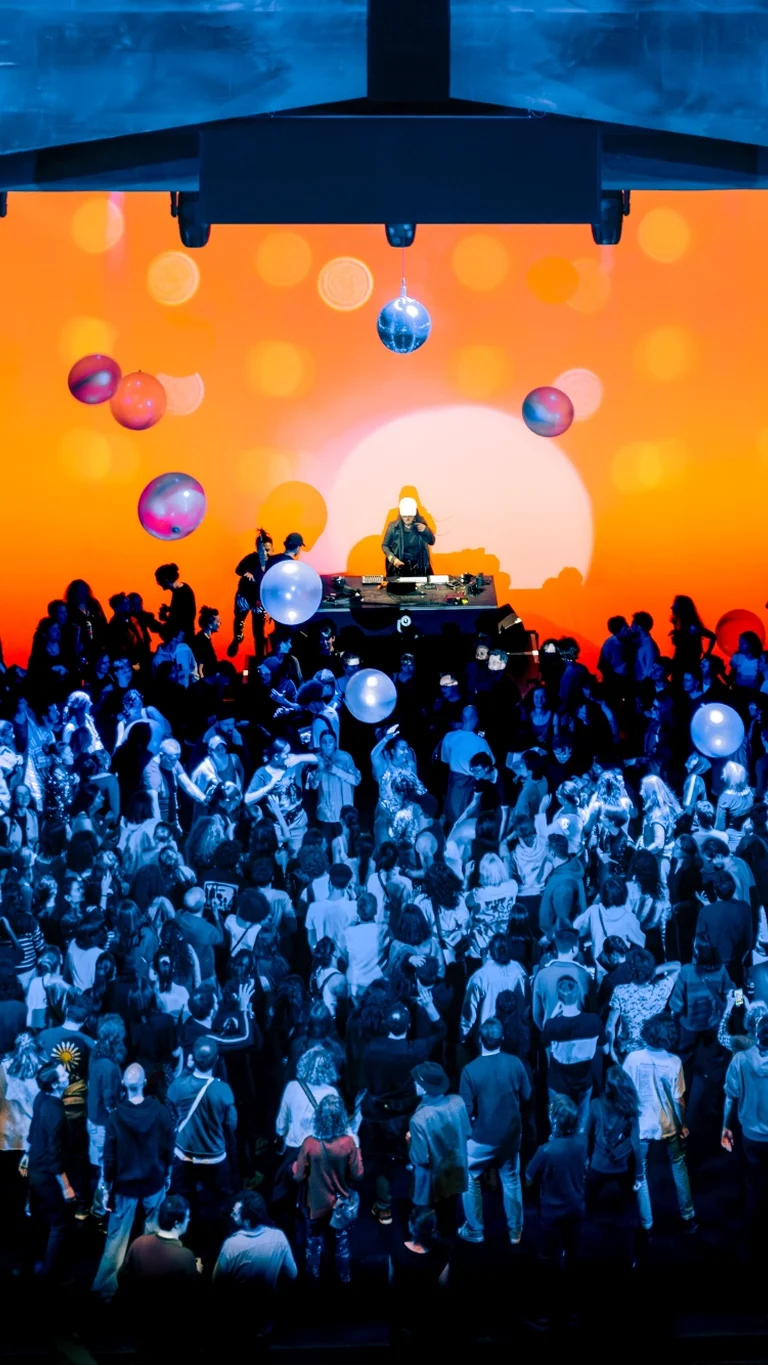 Eine große Menschenmenge tanzt vor einer leuchtend orangefarbenen Bühnenprojektion mit Discokugeln und Ballons, während ein DJ auf einem Podest auflegt.
