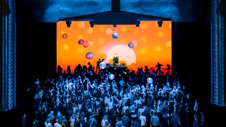 Eine große Menschenmenge tanzt vor einer leuchtend orangefarbenen Bühnenprojektion mit Discokugeln und Ballons, während ein DJ auf einem Podest auflegt.