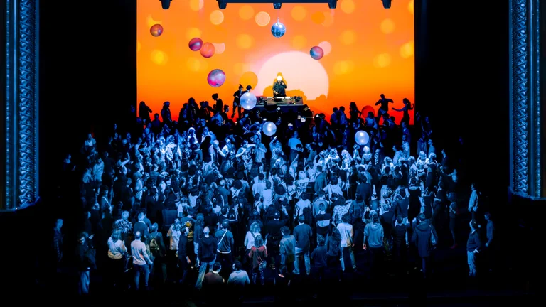 A large crowd dances in front of a brightly orange stage projection with disco balls and balloons, while a DJ performs on a raised platform.