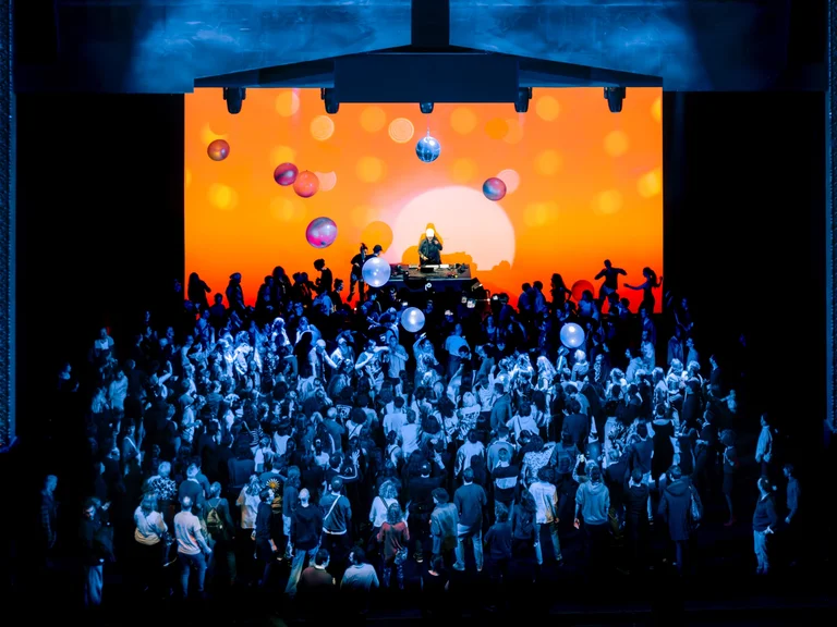 Eine große Menschenmenge tanzt vor einer leuchtend orangefarbenen Bühnenprojektion mit Discokugeln und Ballons, während ein DJ auf einem Podest auflegt.