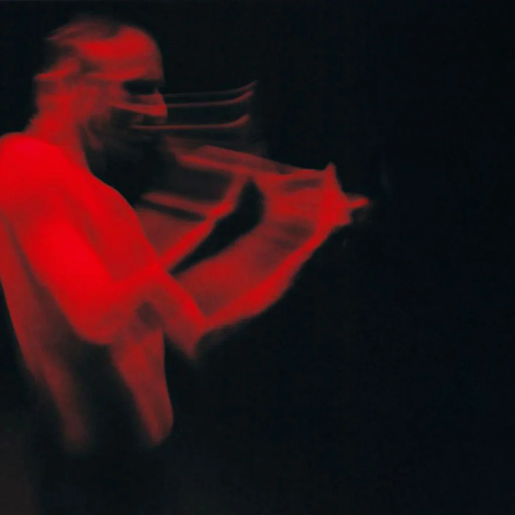 Ein in rotes Licht getauchter, oberkörperfreier Performer. Durch die Bewegungsunschärfe wirkt die Szene dynamisch und fast geisterhaft. Der Hintergrund ist vollständig schwarz.