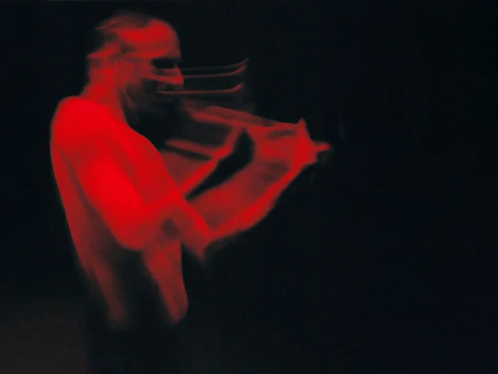 A shirtless musician bathed in red light. The motion blur creates a dynamic and almost ghostly effect. The background is completely black.