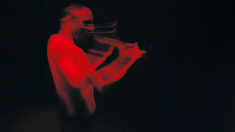 Ein in rotes Licht getauchter, oberkörperfreier Performer. Durch die Bewegungsunschärfe wirkt die Szene dynamisch und fast geisterhaft. Der Hintergrund ist vollständig schwarz.