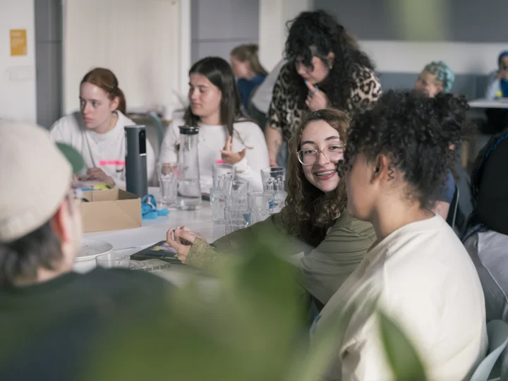 Jugendliche sitzen gemeinsam an einem Tisch und unterhalten sich; im Mittelpunkt lächelt eine junge Person mit Brille und Zahnspange ihrem Gegenüber zu.