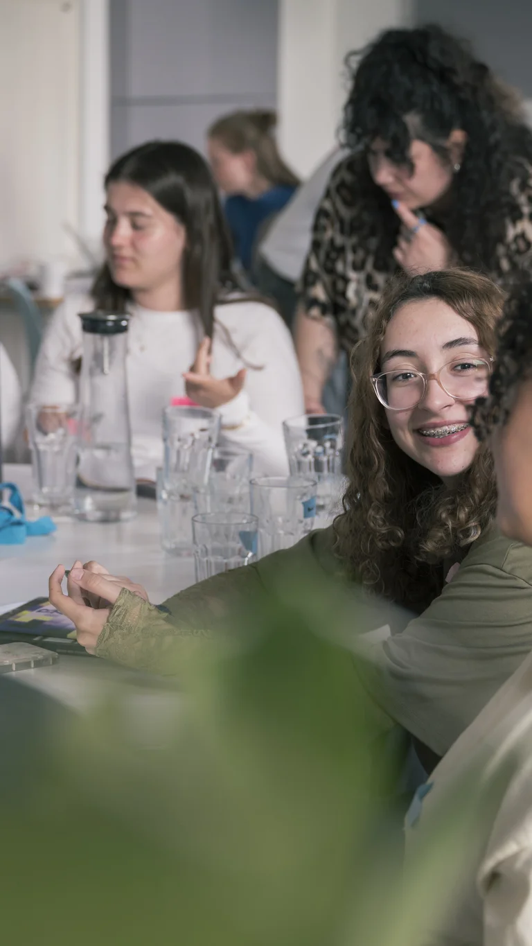 Jugendliche sitzen gemeinsam an einem Tisch und unterhalten sich; im Mittelpunkt lächelt eine junge Person mit Brille und Zahnspange ihrem Gegenüber zu.