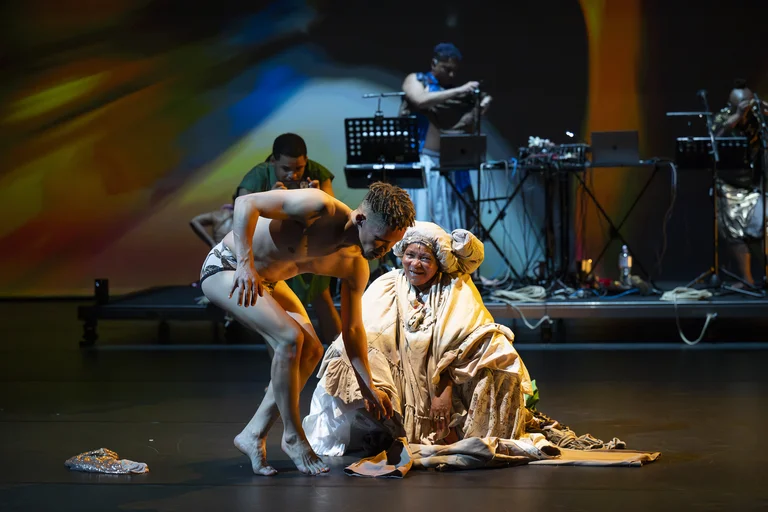 A nearly nude performer in underwear bends toward a smiling actress in elaborate costume, with musicians and projections in the background.