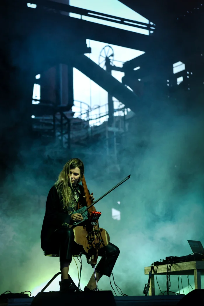 Hildur Guðnadóttir sits on a chair playing an electric string instrument with a bow. Electronic devices and cables are placed beside her. Dense fog and the silhouette of an industrial structure form the backdrop of the stage.