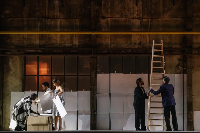 Five performers stand on an elevated stage in front of the window facade of a former industrial hall. On the left, Claron McFadden is bent over a box, while Michael Wilmering and Nina van Essen hold large white sheets of paper. On the right side of the image, Alex Rosen and Steven van Linden are holding a tall wooden ladder.