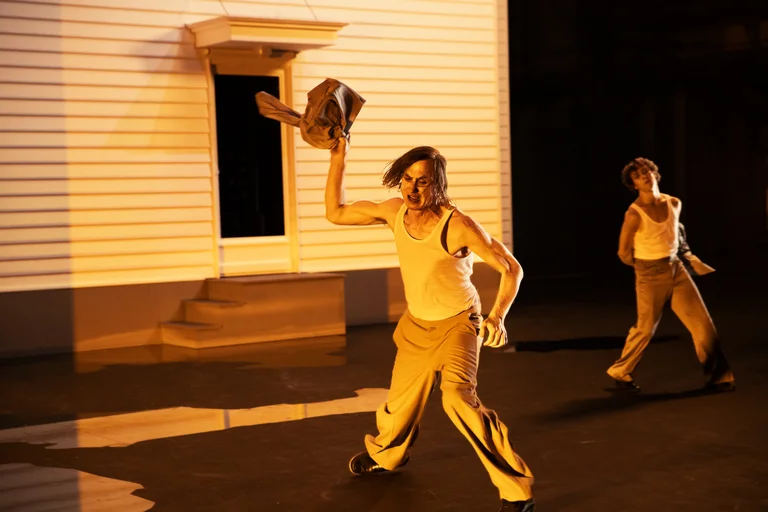 Lars Eidinger dances on a theatre stage in front of a bright house façade. With an expressive look on his face, he energetically throws a piece of clothing into the air. He is wearing a white undershirt and beige trousers. In the background, Samuel Planas can be seen, also wearing a white undershirt, moving with a coat in his hand.