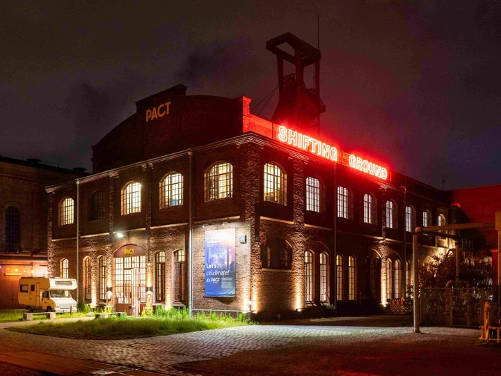 Außenansicht von PACT Zollverein bei Nacht. Auf dem Dach ist eine rot leuchtende Installation mit der Aufschrift "Shifting Ground" zu lesen.