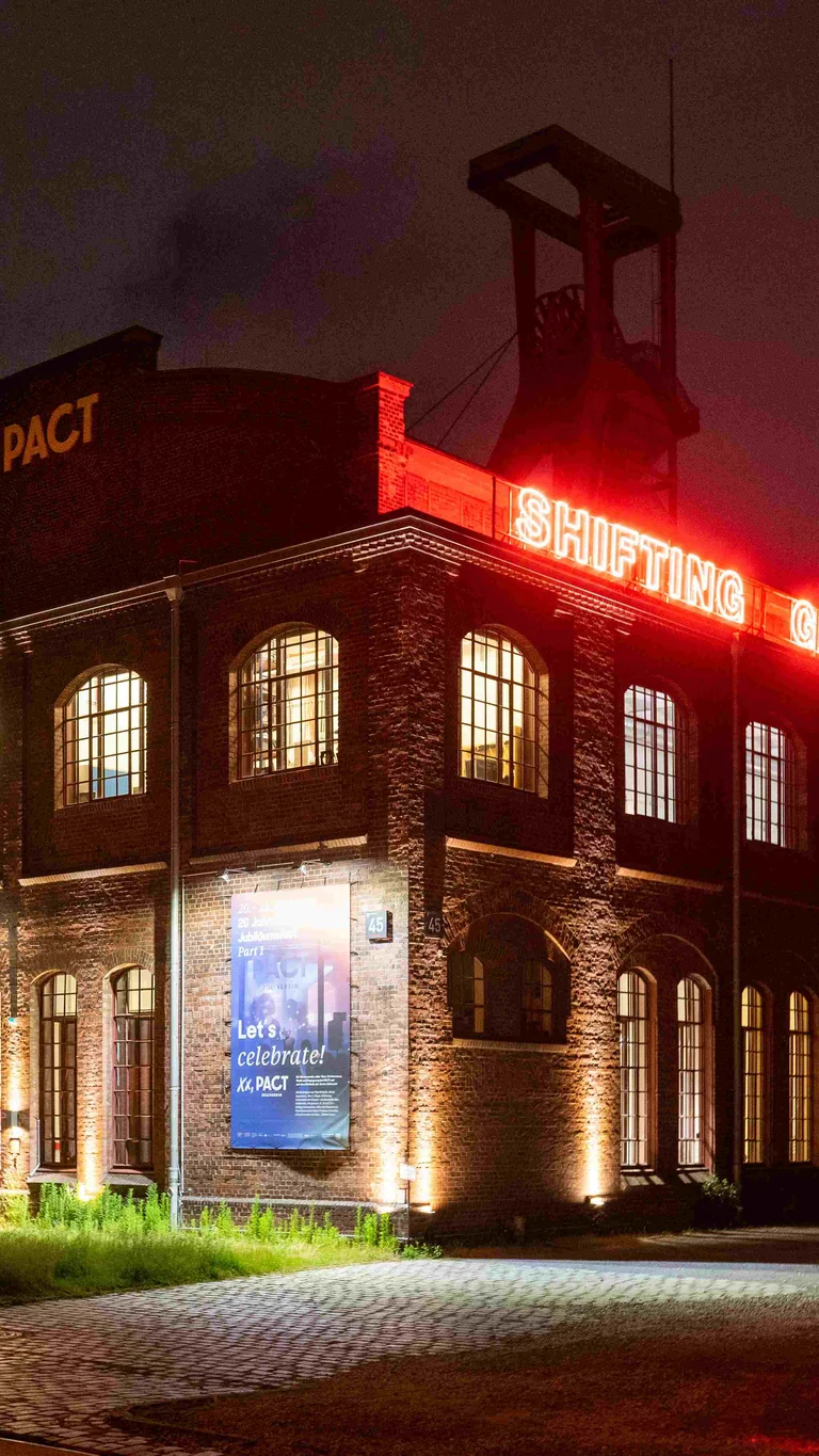Außenansicht von PACT Zollverein bei Nacht. Auf dem Dach ist eine rot leuchtende Installation mit der Aufschrift "Shifting Ground" zu lesen.