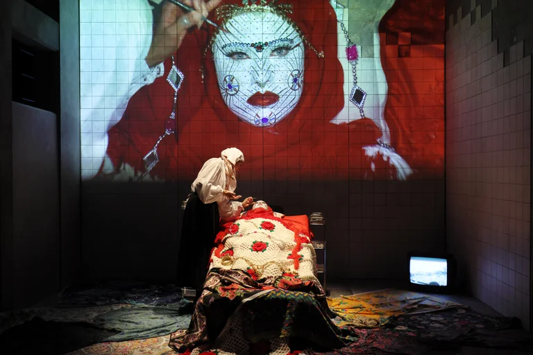 Tiled stage room: one person lies under a colorful embroidered blanket as another leans over applying makeup; above them, a giant projection of a red-veiled face.