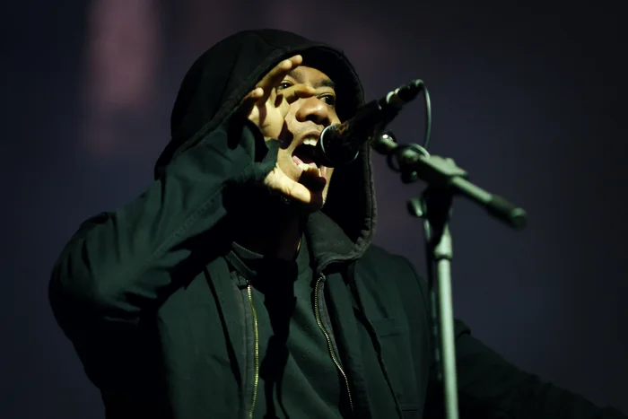 Rully Shabara wears a hooded jacket and sings powerfully into a microphone, emphasizing his voice with his hand. The background is dark.