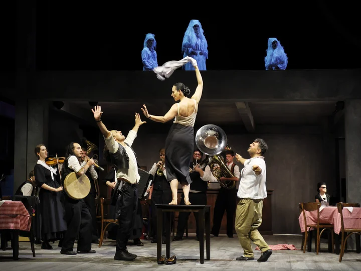 Scene from Balkan Erotic Epic: A dancer stands on a table, waving a cloth, surrounded by performers and musicians in a lively, ritual-like scene. Several white-clad figures appear on an upper level.