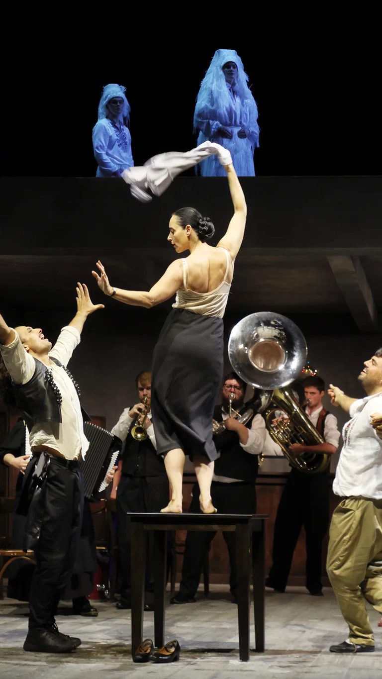 Szene aus Balkan Erotic Epic: Eine Tänzerin steht auf einem Tisch und schwingt ein Tuch, während um sie herum ein Ensemble aus Musiker:innen und Darsteller:innen eine lebhafte, rituelle Szene aufführt. Auf einer oberen Ebene stehen mehrere weiß verhüllte Figuren.