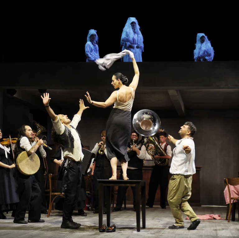 Szene aus Balkan Erotic Epic: Eine Tänzerin steht auf einem Tisch und schwingt ein Tuch, während um sie herum ein Ensemble aus Musiker:innen und Darsteller:innen eine lebhafte, rituelle Szene aufführt. Auf einer oberen Ebene stehen mehrere weiß verhüllte Figuren.