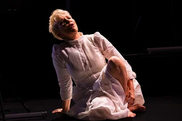 Joyce DiDonato sits barefoot on the dark stage, wearing a white dress. She supports herself with one hand on the floor, one leg bent, and looks upward with her mouth open.