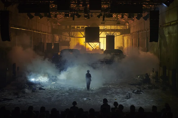 Eine postapokalyptisch wirkende Bühnenszene in einer verlassenen Industriehalle. Dichter Rauch und orangefarbenes Licht erzeugen eine bedrohliche, brennende Atmosphäre. Eine einzelne Gestalt tritt aus dem Nebel hervor, verstreute Trümmer sind im Hintergrund zu erkennen.